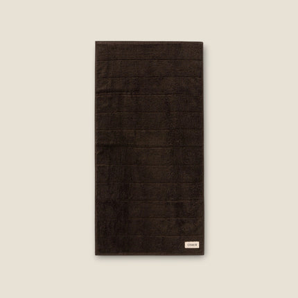 Madhome.ro dark brown cotton bath towel with a soft, dense texture and a small brand label at the bottom.