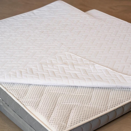 Protectie saltea alba matlasata Madhome.ro din microfibra, cu model geometric, asezata pe o saltea gri.